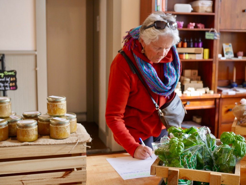 A shopper in a red sweater signs a sheet at a market counter beside jars of sauerkraut and bags of fresh basil